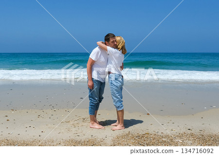 Couple standing barefoot on the beach, embracing near the waves with clear sky and casual outfits. Couple standing barefoot on the beach, embracing near the waves with clear sky and casual outfits. 134716092