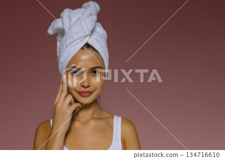 Young woman applying skincare cream to her cheek after a shower, wearing a towel on her head and showing a gentle beauty routine. 134716610