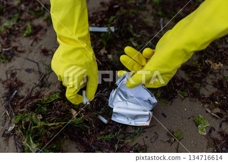 Hands in yellow gloves picking up crushed packaging and litter during beach cleanup, showing pollution, environmental damage and the importance of eco awareness and coastal protection. Hands in yellow gloves picking up crushed packaging and litter during beach cleanup, showing pollution, environmental damage and the importance of eco awareness and coastal protection. 134716614