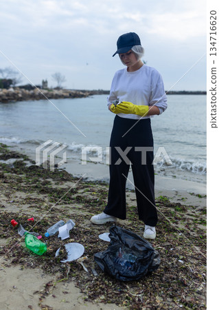 volunteer cleaning polluted beach, collecting plastic bottles, cups and debris in a black bag, supporting ecology, environment protection, sustainability and coastal cleanup awareness. 134716620