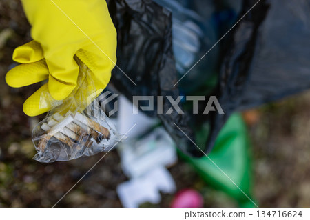 Close-up of a volunteer wearing yellow gloves collecting cigarette butts on a polluted beach. Environmental cleanup, ecology protection, shoreline waste and anti-litter awareness. Close-up of a volunteer wearing yellow gloves collecting cigarette butts on a polluted beach. Environmental cleanup, ecology protection, shoreline waste and anti-litter awareness. 134716624