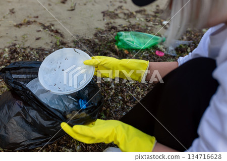 Volunteer collecting plastic waste and litter into a black trash bag during beach cleanup, highlighting pollution, environmental protection, sustainability, and eco awareness. Volunteer collecting plastic waste and litter into a black trash bag during beach cleanup, highlighting pollution, environmental protection, sustainability, and eco awareness. 134716628
