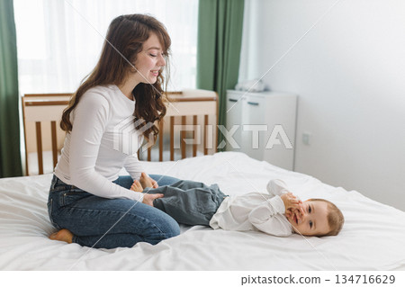 Young mother playing with her baby on a soft white bed, enjoying a warm and cozy family moment full of comfort, tenderness, joy, and natural home light. 134716629