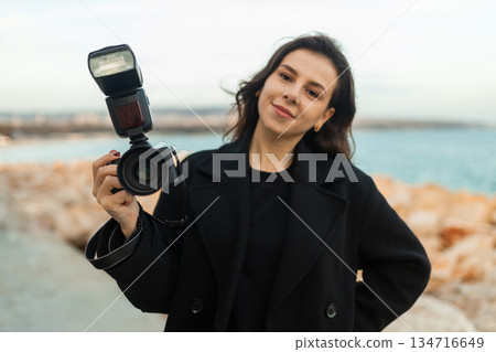 Young female photographer taking pictures outdoors by the sea, smiling while holding a professional camera with flash, capturing moments on a rocky coastline at sunset. Young female photographer taking pictures outdoors by the sea, smiling while holding a professional camera with flash, capturing moments on a rocky coastline at sunset. 134716649