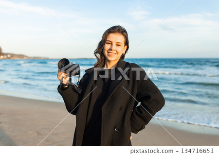 Smiling female photographer holding a camera on a sunny beach, enjoying golden hour light and capturing outdoor moments on the coastline. 134716651