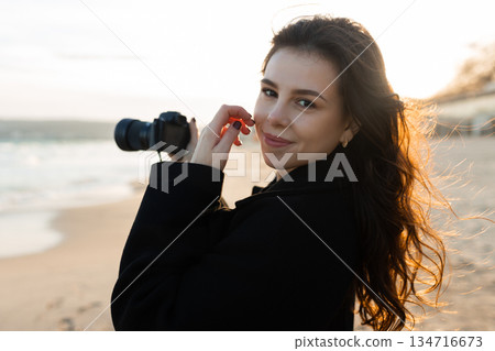 Smiling female photographer holding a camera on a sunny beach, enjoying golden hour light and capturing outdoor moments on the coastline. 134716673