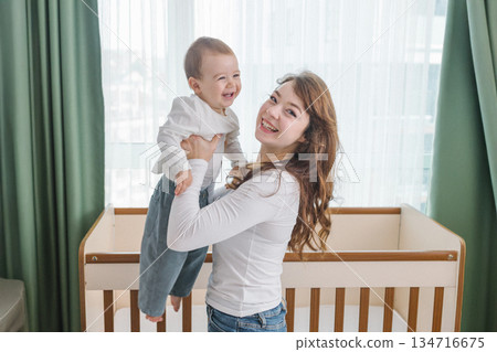 Warm and cozy family moment of a young mother lifting her smiling baby in a bright home interior, enjoying joy, comfort, natural light, and playful parent-child bonding. 134716675