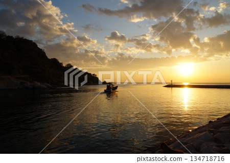 A colorful wooden boat sets out to sea at dawn to fish. It's morning, dawn, and hot. In the background is a mountain with green trees and grass and a blue sky.golden dawn on the seashore A colorful wooden boat sets out to sea at dawn to fish. It's morning, dawn, and hot. In the background is a mountain with green trees and grass and a blue sky.golden dawn on the seashore 134718716