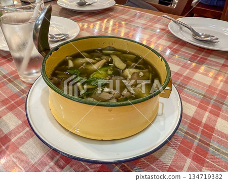 A clear herbal soup with vegetables and meat sits in a small enamel pot. The dish is served hot as a shared meal on a dining table. 134719382