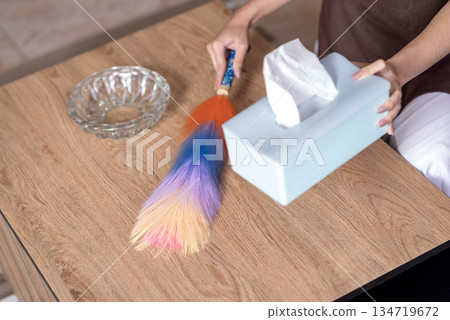 Closeup of a woman using a feather duster to clean a wooden table, with a tissue box and glass ashtray nearby. Hygiene and cleanliness Closeup of a woman using a feather duster to clean a wooden table, with a tissue box and glass ashtray nearby. Hygiene and cleanliness 134719672
