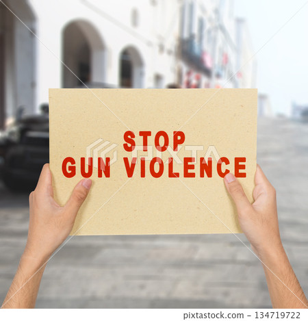 Hands holding a simple sign with the message of Stop Gun Violence written in red letters, with a blurred city street in the background. Concept of gun violence 134719722