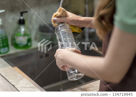 Person hands washing a clear plastic bottle with a yellow sponge under a stream of water in a kitchen sink, promoting cleanliness and hygiene Person hands washing a clear plastic bottle with a yellow sponge under a stream of water in a kitchen sink, promoting cleanliness and hygiene 134719945