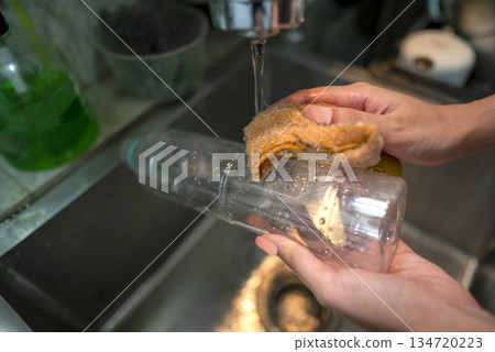 Person hands washing a clear plastic bottle with a yellow sponge under a stream of water in a kitchen sink, promoting cleanliness and hygiene Person hands washing a clear plastic bottle with a yellow sponge under a stream of water in a kitchen sink, promoting cleanliness and hygiene 134720223