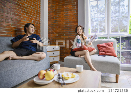 Indonesian southeast asian couple is relaxing together. The man is playing a game on his cellphone, while the woman is listening to music with her headphones and reading a book 134721286