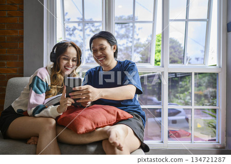 Indonesian southeast asian couple is sitting together on a couch, while looking at something on a cellphone. They are both smiling and enjoying, creating a warm and relatable atmosphere Indonesian southeast asian couple is sitting together on a couch, while looking at something on a cellphone. They are both smiling and enjoying, creating a warm and relatable atmosphere 134721287