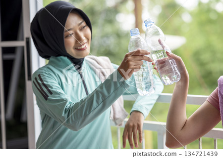 Two Indonesian southeast asian women wearing sportswear and a towel around neck is holding a water bottle after a workout. She appears refreshed and healthy 134721439