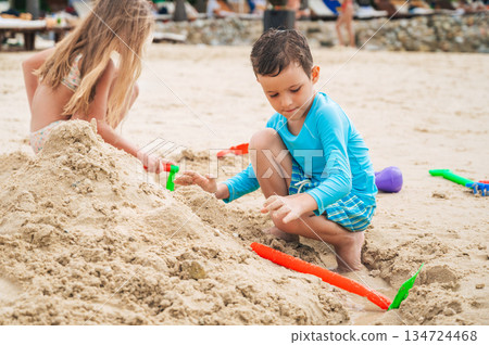 Cute child boy plays on beach and builds sand castle with friends in summer by the sea on vacation 134724468