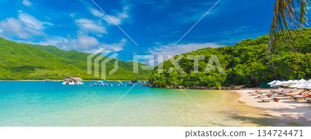 landscape panorama with sandy beach with sun loungers and umbrellas in blue lagoon by the sea on a sunny summer day at a resort on paradise island 134724471