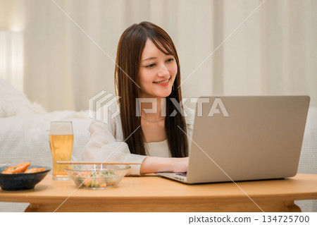 Young Asian woman relaxing and looking at laptop while eating at home in the evening Young Asian woman relaxing and looking at laptop while eating at home in the evening 134725700