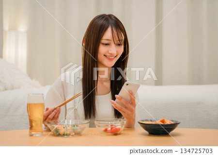 一位年輕的亞洲女性在家裡的臥室裡一邊喝啤酒一邊看智慧型手機(啤酒,在家喝酒,飲酒習慣) 一位年輕的亞洲女性在家裡的臥室裡一邊喝啤酒一邊看智慧型手機(啤酒,在家喝酒,飲酒習慣) 134725703
