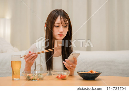 一位年輕的亞洲女性在家裡的臥室裡一邊喝啤酒一邊看智慧型手機(啤酒,在家喝酒,飲酒習慣) 一位年輕的亞洲女性在家裡的臥室裡一邊喝啤酒一邊看智慧型手機(啤酒,在家喝酒,飲酒習慣) 134725704