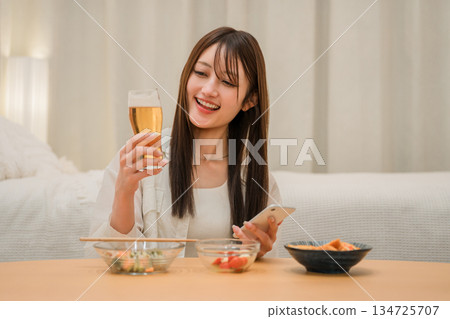 一位年輕的亞洲女性在家裡的臥室裡一邊喝啤酒一邊看智慧型手機(啤酒,在家喝酒,飲酒習慣) 一位年輕的亞洲女性在家裡的臥室裡一邊喝啤酒一邊看智慧型手機(啤酒,在家喝酒,飲酒習慣) 134725707