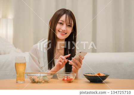 一位年輕的亞洲女性在家裡的臥室裡一邊喝啤酒一邊看智慧型手機(啤酒,在家喝酒,飲酒習慣) 一位年輕的亞洲女性在家裡的臥室裡一邊喝啤酒一邊看智慧型手機(啤酒,在家喝酒,飲酒習慣) 134725710