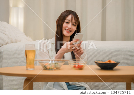 一位年輕的亞洲女性在家裡的臥室裡一邊喝啤酒一邊看智慧型手機(啤酒,在家喝酒,飲酒習慣) 一位年輕的亞洲女性在家裡的臥室裡一邊喝啤酒一邊看智慧型手機(啤酒,在家喝酒,飲酒習慣) 134725711