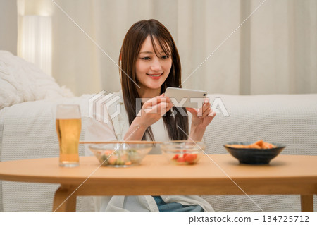 一位年輕的亞洲女性在家裡的臥室裡一邊喝啤酒一邊看智慧型手機(啤酒,在家喝酒,飲酒習慣) 一位年輕的亞洲女性在家裡的臥室裡一邊喝啤酒一邊看智慧型手機(啤酒,在家喝酒,飲酒習慣) 134725712