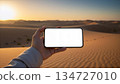 Mockup image of a hand holding mobile phone with blank screen in the middle of the desert 134727010