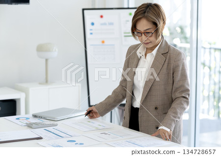 Senior female executive is standing looking at documents and computers arranged Senior female executive is standing looking at documents and computers arranged 134728657