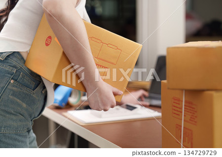 E-commerce. Woman carrying package while managing online business in home office. 134729729