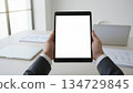 Tablet with a blank screen held in a modern office. Concept of business, remote work, digital productivity, technology, and professional workplace. Ideal for presentations, advertising, and stock illu 134729845
