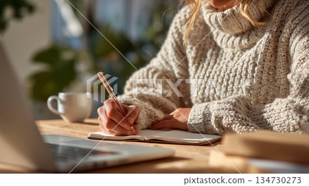 Cozy woman writing in notebook at home workspace with laptop and coffee Cozy woman writing in notebook at home workspace with laptop and coffee 134730273