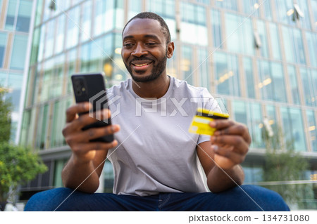 African man shopping online holding smartphone paying with gold credit card. Guy on urban street in city buying on Internet enter credit card details. Online shopping ecommerce delivery service African man shopping online holding smartphone paying with gold credit card. Guy on urban street in city buying on Internet enter credit card details. Online shopping ecommerce delivery service 134731080