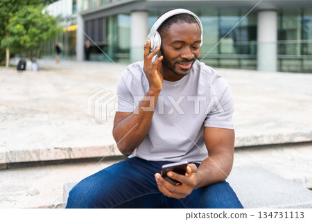 Music song concept. Good start day. Happy African American man listening modern hit on street in city. Person guy wearing headphones enjoy listening favorite music on phone. People lifestyle joy Music song concept. Good start day. Happy African American man listening modern hit on street in city. Person guy wearing headphones enjoy listening favorite music on phone. People lifestyle joy 134731113