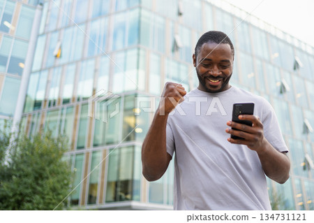 Happy African American man euphoric winner with smartphone on street in city. Person guy looking at cell phone reading great news getting good result winning online bid feeling amazed. Winning gesture Happy African American man euphoric winner with smartphone on street in city. Person guy looking at cell phone reading great news getting good result winning online bid feeling amazed. Winning gesture 134731121
