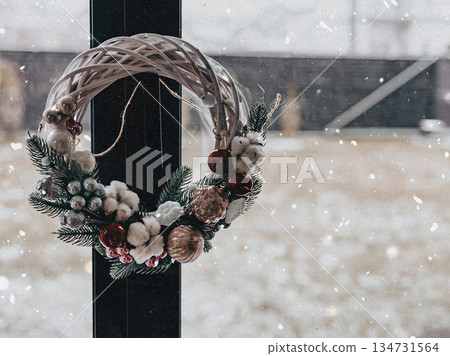 Cozy winter holiday scene. Handmade Christmas wreath hanging on a window pane with snow falling outdoors, festive mood, domestic life, seasonal warmth 134731564