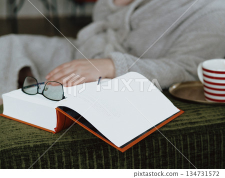 Quiet winter moment. Unrecognizable person relaxing with a paper book, hot coffee on a green corduroy sofa at cozy home. A middle-aged woman relaxing. 134731572