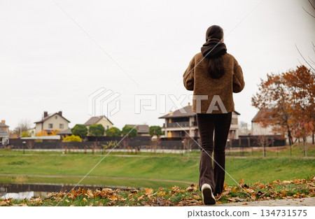 Stylish young woman walking outdoors in autumn park with smartphone, cozy brown outfit, calm mindful lifestyle, slow living female lifestyle. 134731575