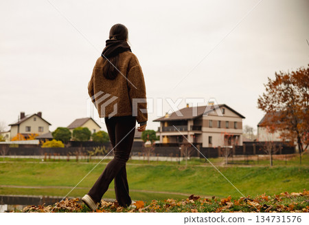 Stylish young woman walking outdoors in autumn park with smartphone, cozy brown outfit, calm mindful lifestyle, slow living female lifestyle. 134731576