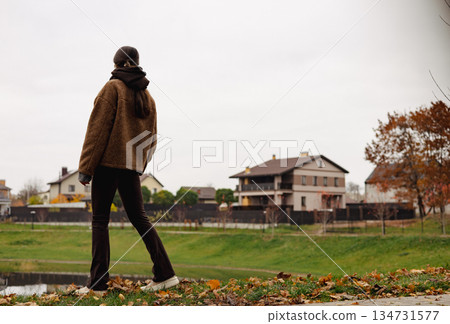 Stylish young woman walking outdoors in autumn park with smartphone, cozy brown outfit, calm mindful lifestyle, slow living female lifestyle. 134731577
