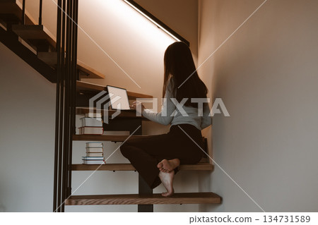 Young woman working on laptop at home office sitting on wooden stairs, cozy minimalist interior, remote work and creative freelance lifestyle concept, calm productivity and modern slow living vibe. 134731589