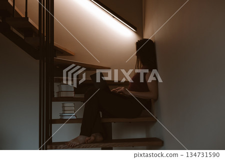 Young woman working on laptop at home office sitting on wooden stairs, cozy minimalist interior, remote work and freelance lifestyle, slow living vibe 134731590