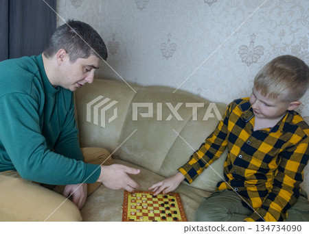 Father teaching his young son to play checkers at home. Warm family moment, parenting, learning through play, quality time together, casual indoor lifestyle scene. Father teaching his young son to play checkers at home. Warm family moment, parenting, learning through play, quality time together, casual indoor lifestyle scene. 134734090
