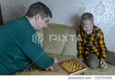 Father teaching his young son to play checkers at home. Warm family moment, parenting, learning through play, quality time together, casual indoor lifestyle scene. Father teaching his young son to play checkers at home. Warm family moment, parenting, learning through play, quality time together, casual indoor lifestyle scene. 134734091