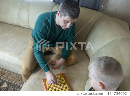 Father teaching his young son to play checkers at home. Warm family moment, parenting, learning through play, quality time together, casual indoor lifestyle scene. 134734092