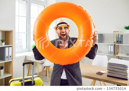 Joyful happy man holding yellow suitcase and swim ring while standing in office 134735014