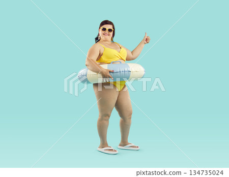 Fat woman in rubber ring wearing swimsuit showing thumb up sign on blue background. 134735024