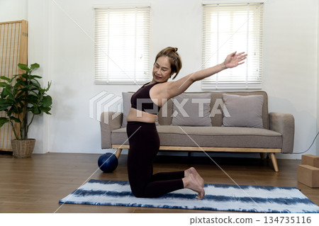Adult woman 50+ in workout clothes kneeling on a mat and stretching 134735116